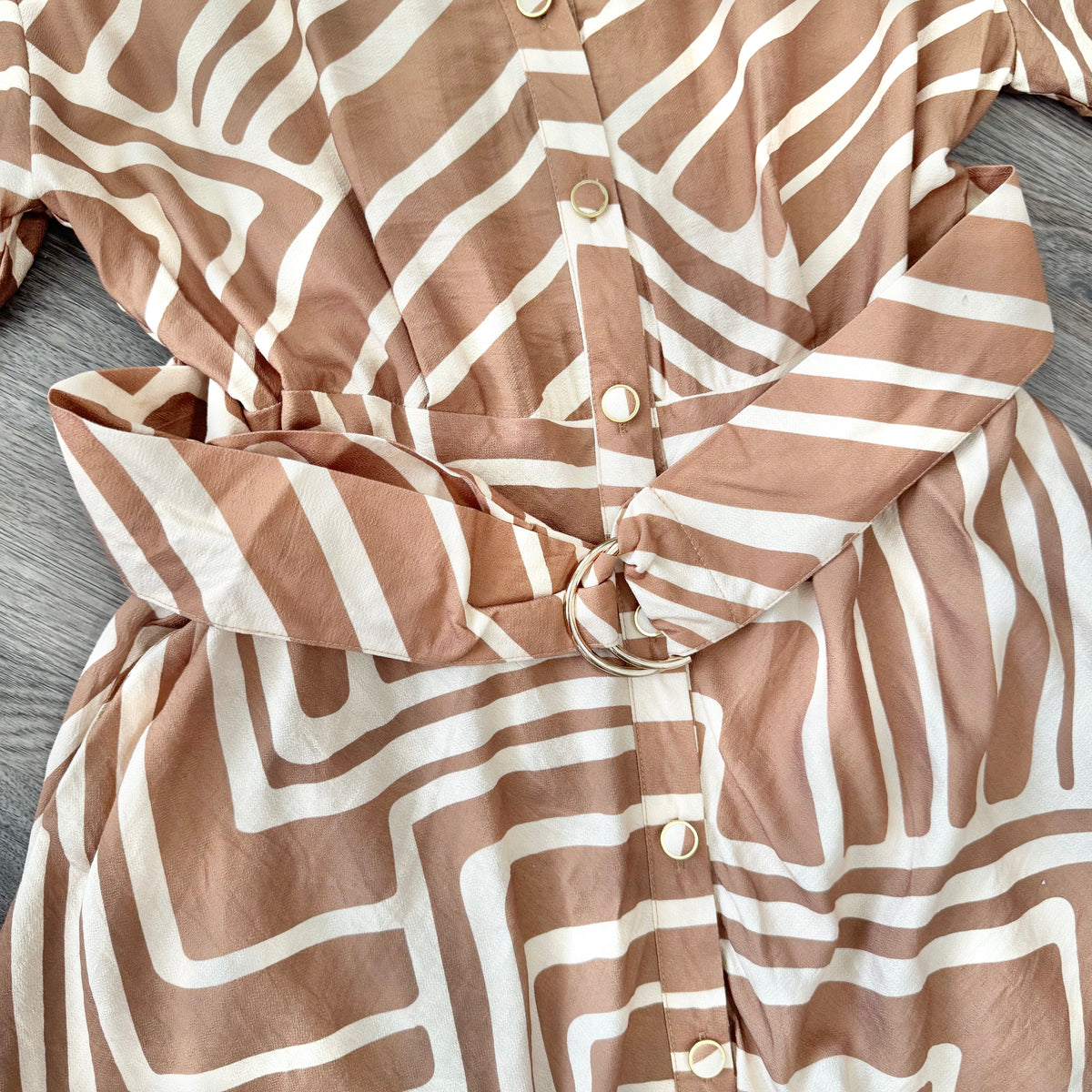 A close-up of a button-front garment with an abstract wavy stripe pattern in warm taupe-brown and off-white, tied at the waist with a matching fabric belt. The garment is laid flat on a gray wood surface.
