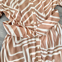 A close-up of a button-front garment with an abstract wavy stripe pattern in warm taupe-brown and off-white, tied at the waist with a matching fabric belt. The garment is laid flat on a gray wood surface.