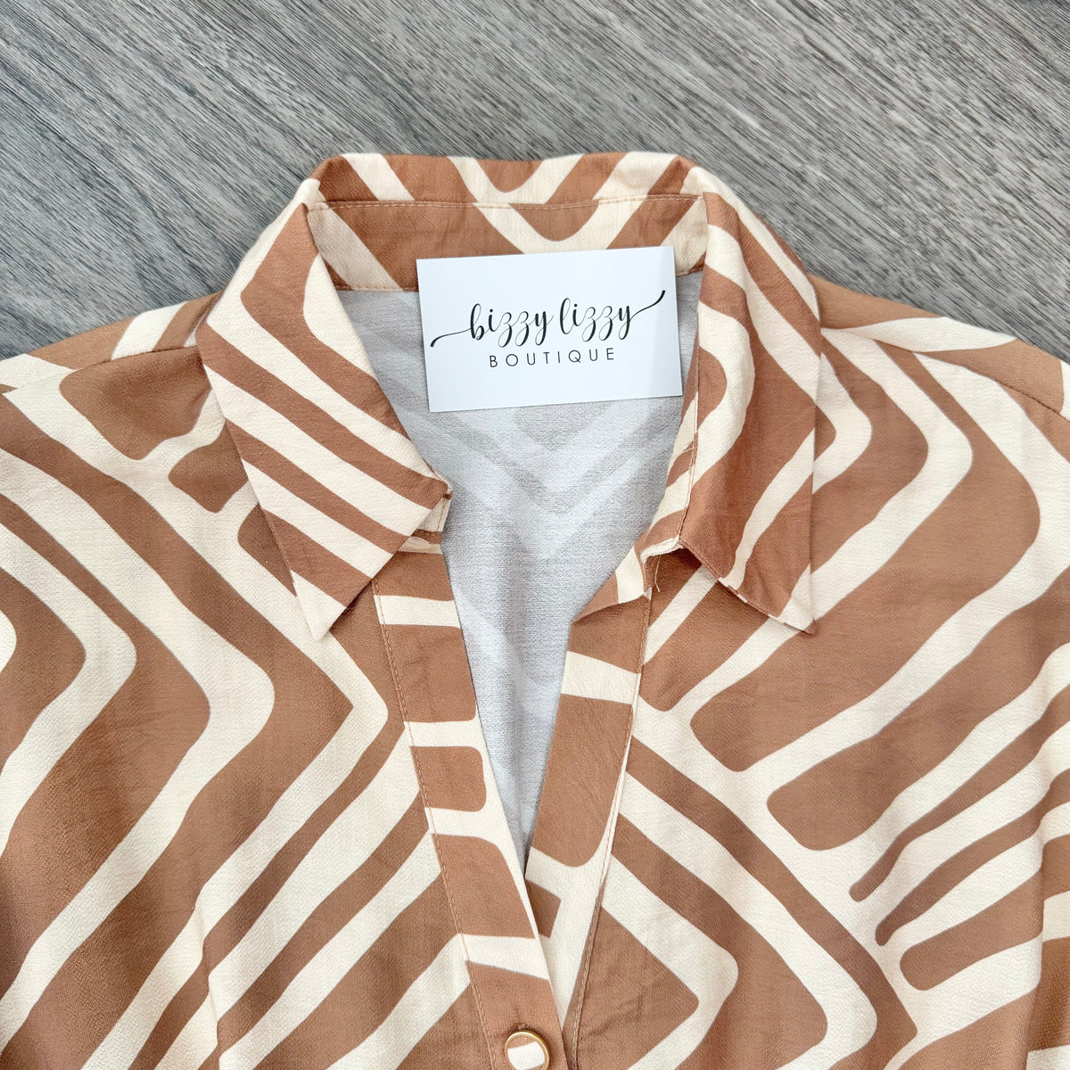 A close-up of a collared button-front blouse laid flat on a gray wood-textured surface. The fabric has a wavy geometric stripe pattern in warm tan and cream, with a visible neck label reading 'bizzy lizzy BOUTIQUE'.