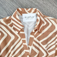 A close-up of a collared button-front blouse laid flat on a gray wood-textured surface. The fabric has a wavy geometric stripe pattern in warm tan and cream, with a visible neck label reading 'bizzy lizzy BOUTIQUE'.