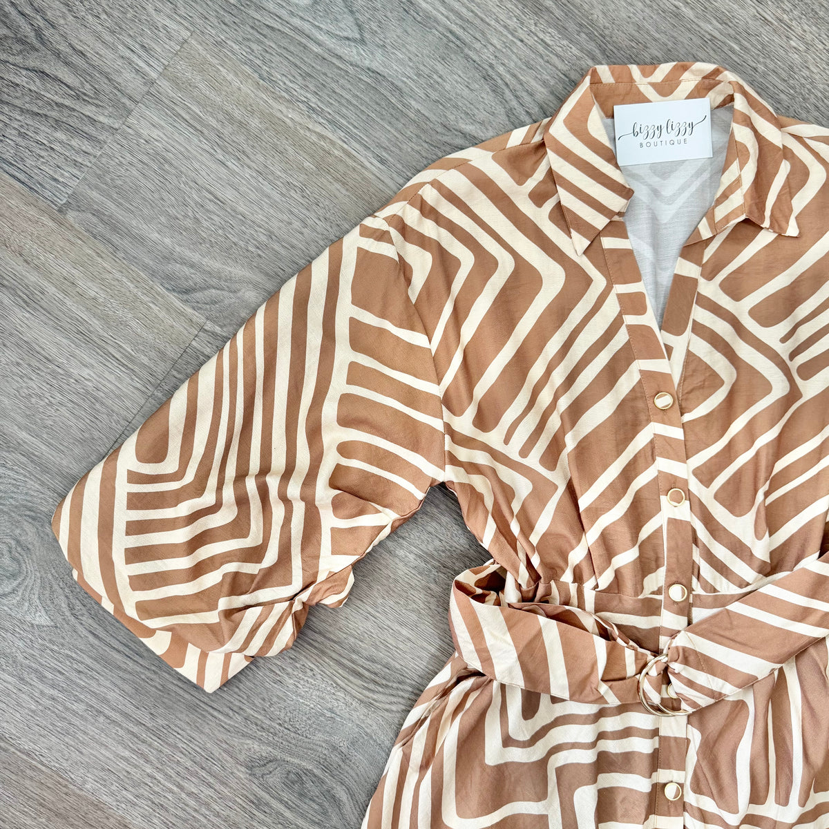 A tan and cream geometric patterned shirt dress with a tie waist, laid flat on a gray wood floor. The brand label reads 'Bizzy Lizzy Boutique'.
