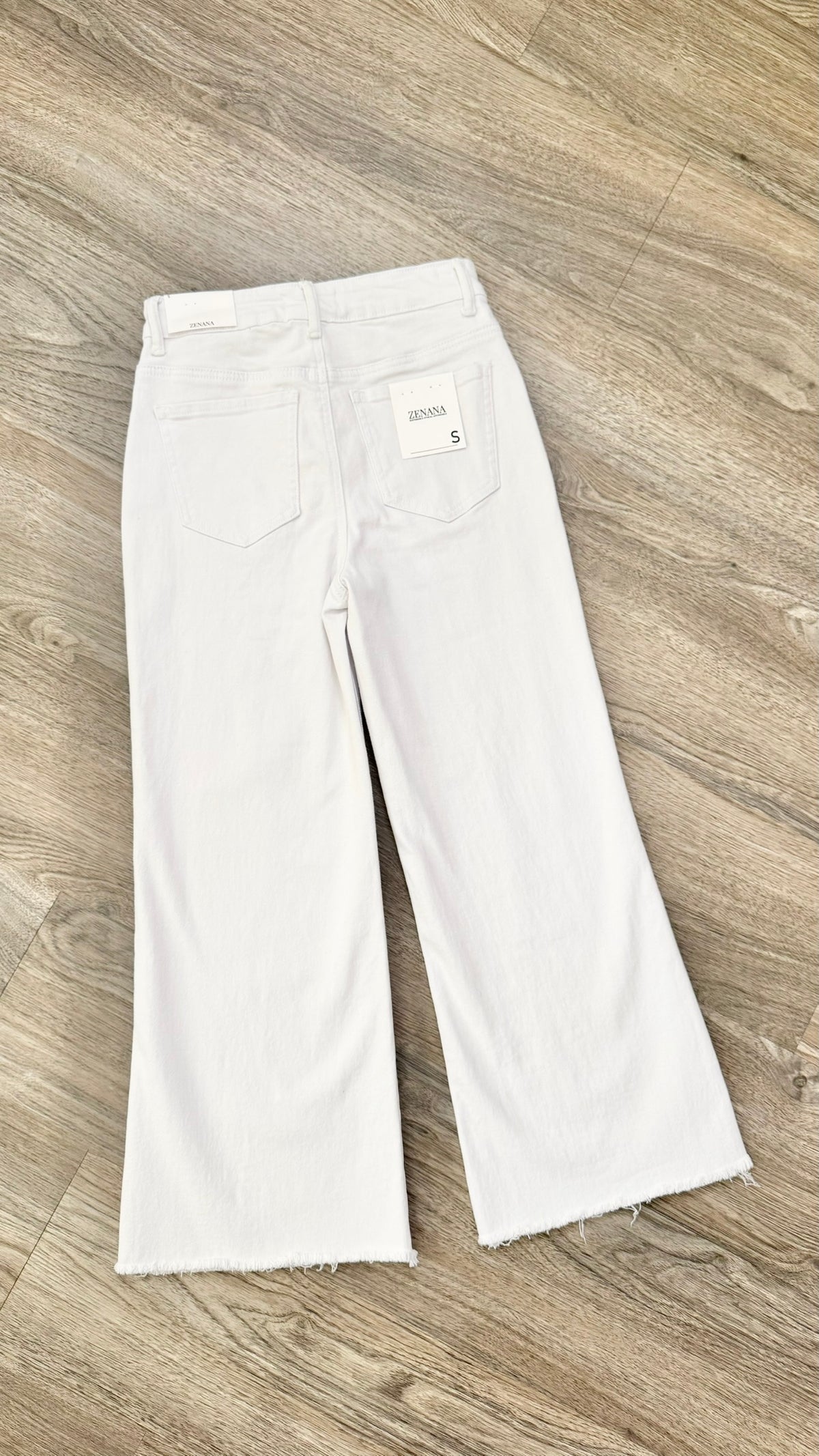 White wide-leg cropped jeans laid flat on a light brown wood floor, showing the back pockets and a small white brand label.