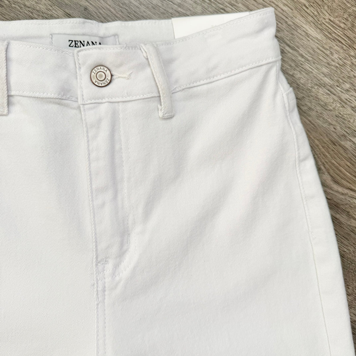 White denim shorts with a silver button closure, laid flat on a gray wood-textured surface. The brand name 'ZENARA' is visible on the inner waistband label.