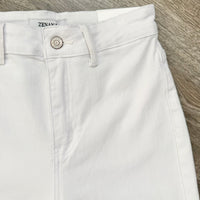White denim shorts with a silver button closure, laid flat on a gray wood-textured surface. The brand name 'ZENARA' is visible on the inner waistband label.