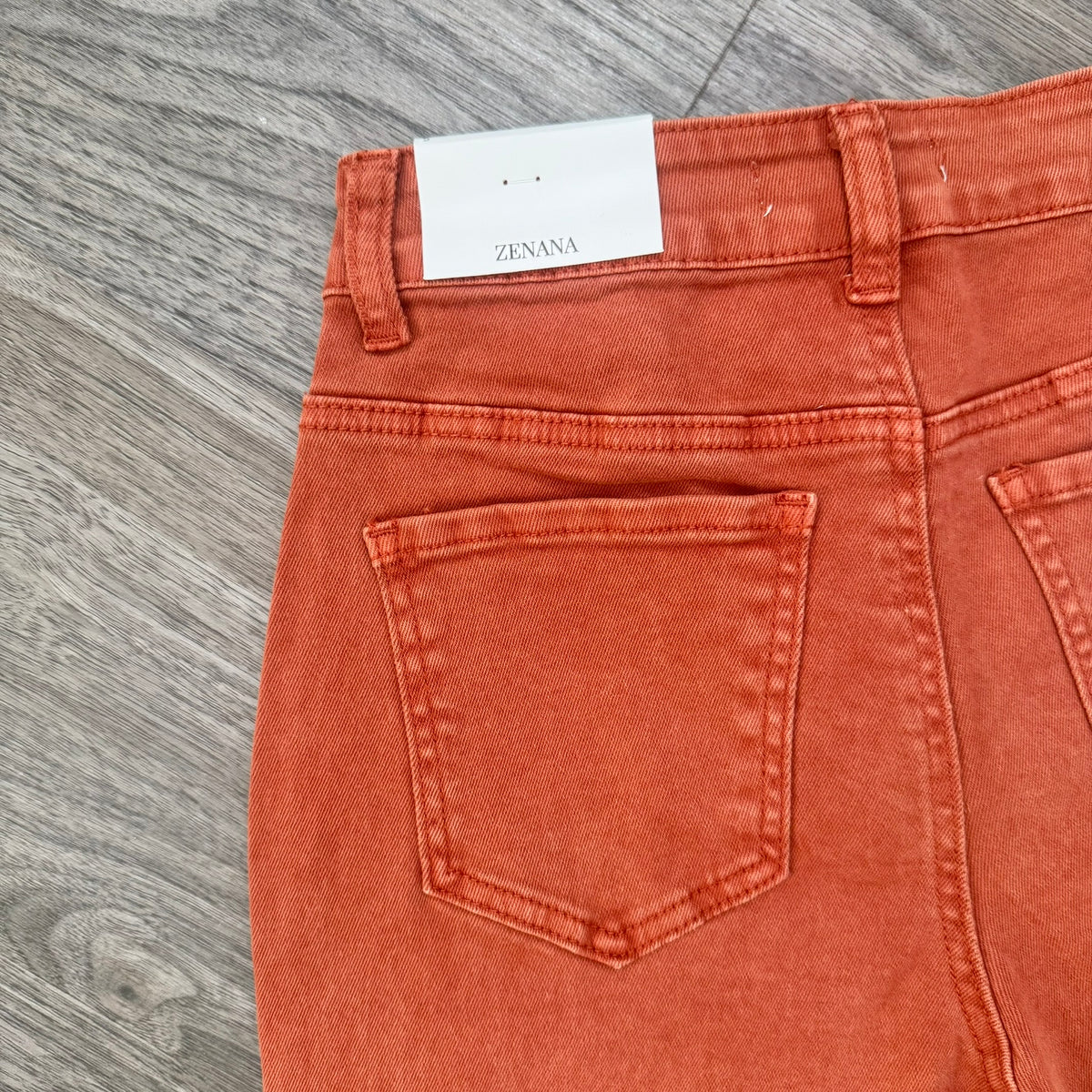 Close-up of the back waistband and rear pockets of orange denim jeans with a white Zenana brand label visible at the waist, laid flat on a gray wood-textured surface.