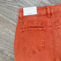 Close-up of the back waistband and rear pockets of orange denim jeans with a white Zenana brand label visible at the waist, laid flat on a gray wood-textured surface.