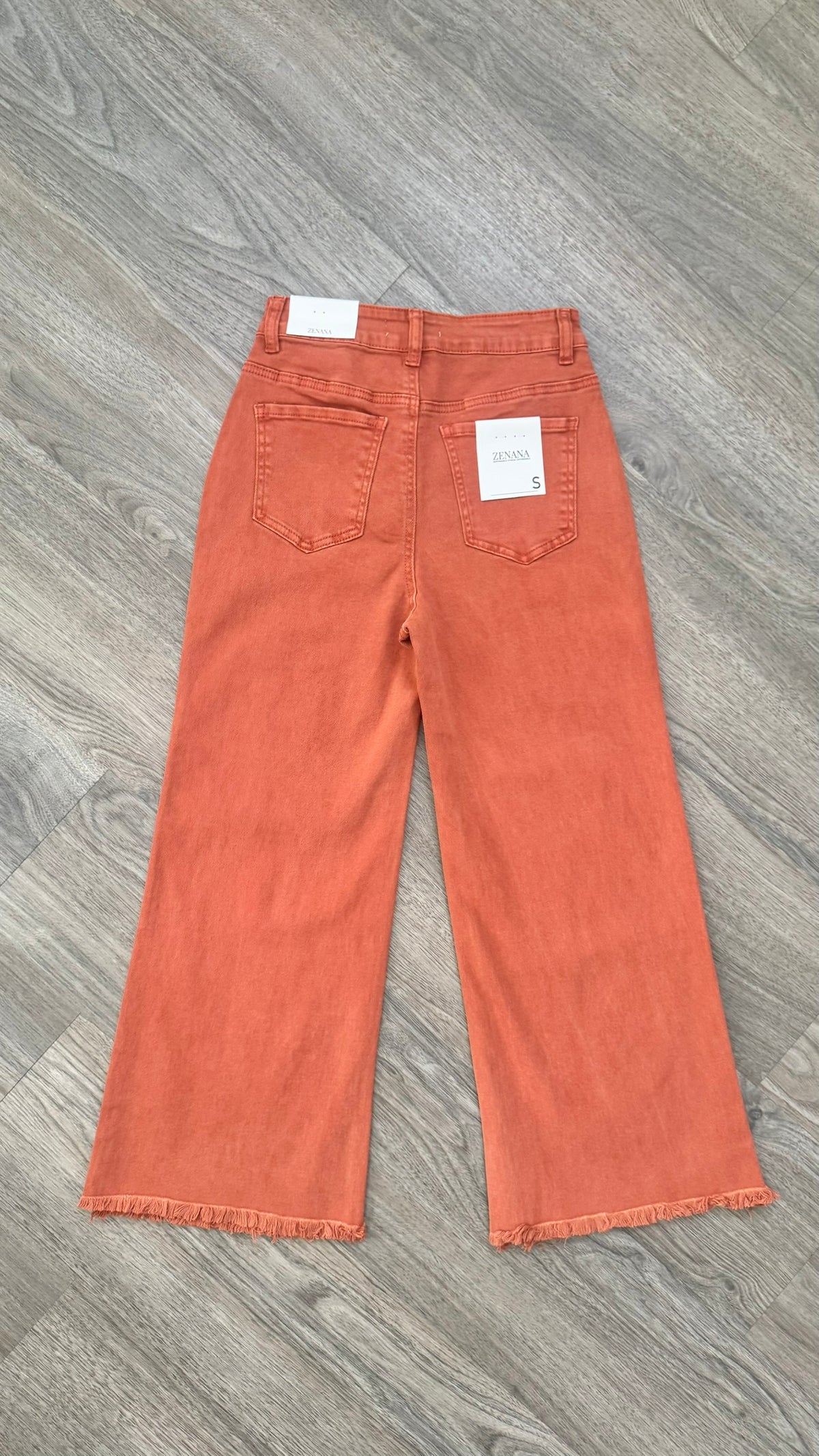 A pair of wide-leg cropped jeans in a faded terracotta or rust orange color, laid flat on a gray wood-look floor. The back pockets and white paper tags are visible.