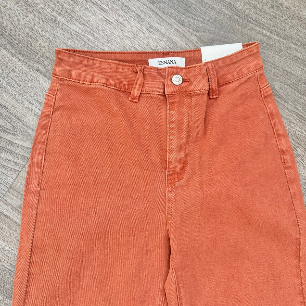 A pair of high-waisted, wide-leg denim pants in a washed terracotta or rust orange color, laid flat on a gray wood-textured surface. 