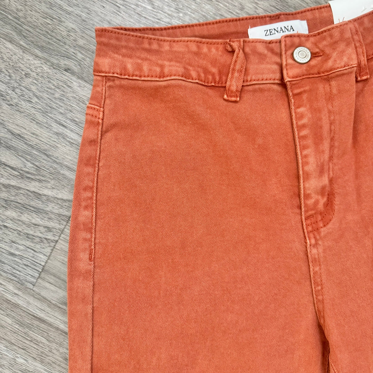 A close-up of a pair of burnt orange denim jeans laid flat on a gray wood-textured surface, showing the waistband, button, belt loops, and front pocket.
